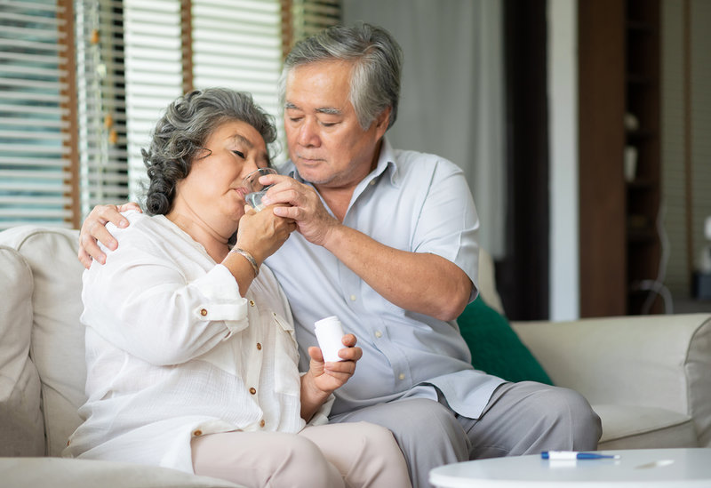 Een grijze, Aziatische man helpt zijn vrouw met het nemen van haar medicijnen